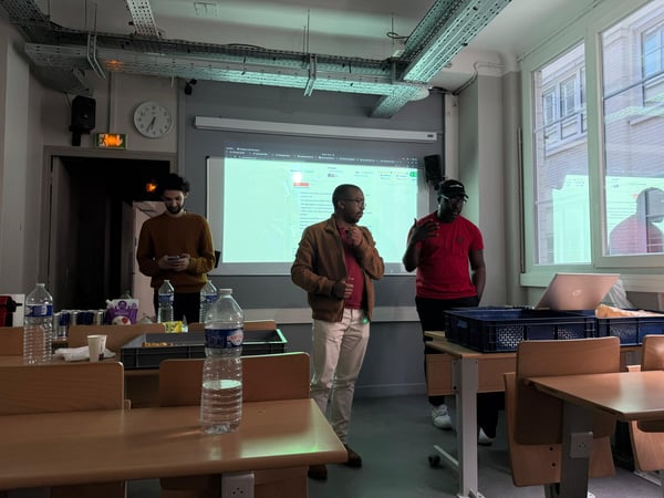 Three men are at the front of a classroom during a presentation, with one man speaking and another gesturing while a projector screen displays a website in the background.