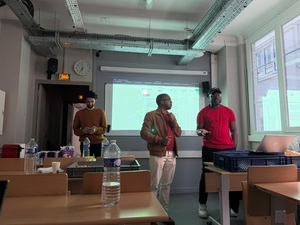 A group of three men are standing in a classroom or office space in front of a projector screen, likely giving a presentation or demonstration.