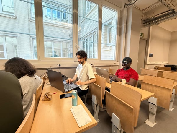 Three people are sitting at wooden desks in a classroom or study area, focused on their laptops.