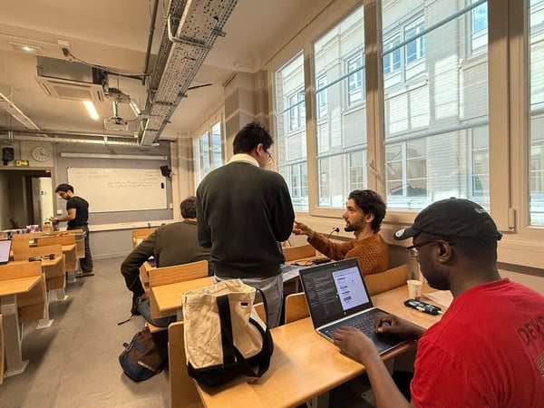A group of young men are gathered in a classroom or workshop setting, with some working on laptops and others engaged in conversation.