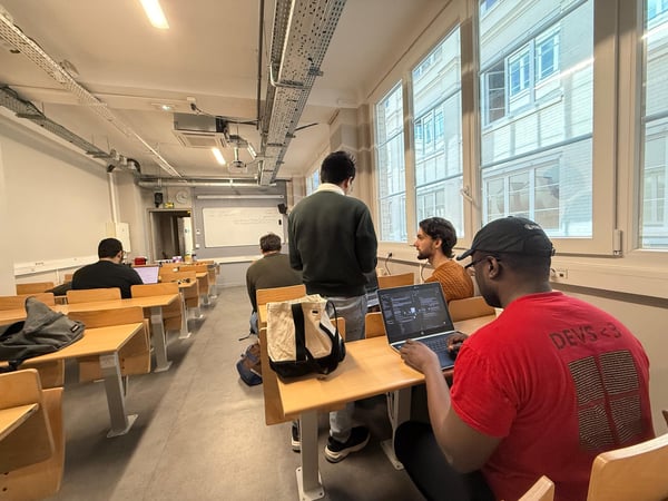 A classroom or workshop setting where several people are working on laptops, with a person in a red shirt in the foreground.