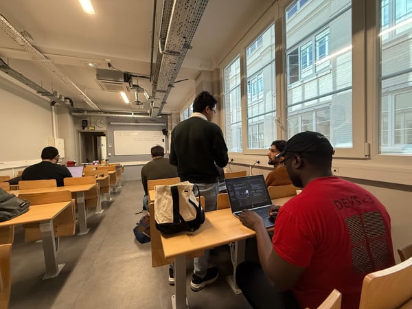 A group of people are gathered in a classroom or lab setting, with individuals sitting at desks using laptops and one person standing while engaging in a discussion.