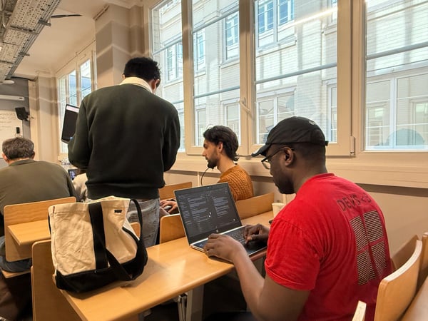 A group of people are working in a classroom or office setting, with one individual in a red shirt actively using a laptop in the foreground.