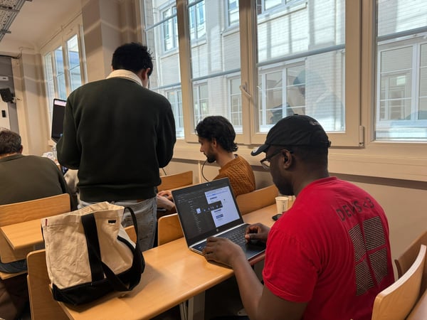 A group of individuals are working on laptops in a classroom or office setting with large windows in the background.