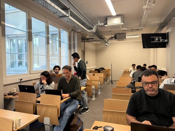 A photograph showing several people working on laptops in a classroom or study hall with large windows and an industrial-style ceiling.