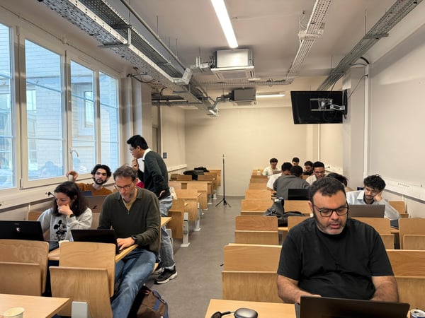 A group of people are seated at wooden desks in a classroom or lecture hall, most of them working on laptops.