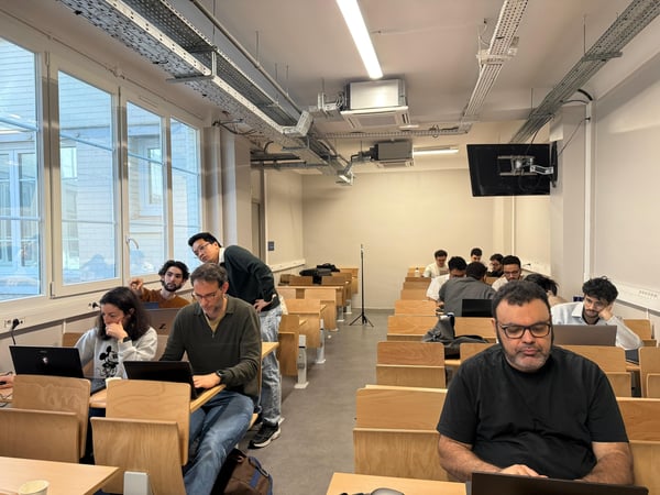 A group of people are seated at wooden desks in a classroom or lecture hall, most of them working on laptops.