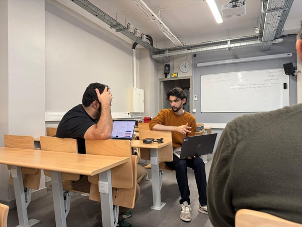 Two men are sitting at desks with laptops in a classroom-like setting, appearing to be in a discussion or working together.