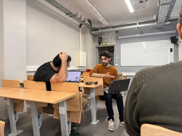 Two men are engaged in a discussion in a classroom or office setting, with one gesturing while holding a laptop and a whiteboard visible in the background.