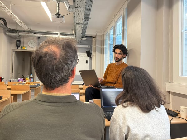 A young man in an orange sweater presents content on a laptop to two individuals sitting in front of him in a classroom or workshop setting.