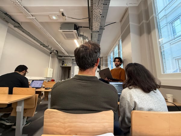 A young man stands at the front of a classroom, presenting from a laptop to a small group of people seated at desks.