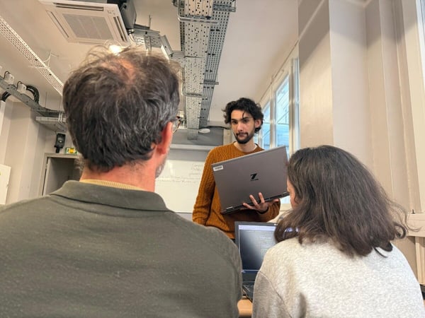 A man in an orange sweater presents a laptop to two colleagues in an office or classroom setting with a whiteboard in the background.