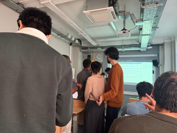 A group of people are gathered in a room, likely a classroom or office, facing a projection screen during a presentation or meeting.