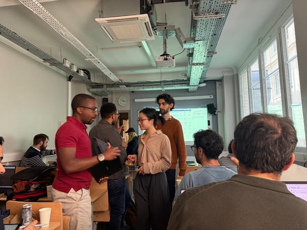 A group of people are gathered in a modern indoor space, likely a classroom or office, engaged in discussion with laptops and a projector screen visible.