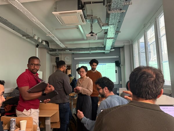 A group of people, likely students or colleagues, are gathered in a classroom or office setting, engaging in a discussion near a whiteboard and laptops.