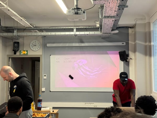 A photo of an indoor classroom or presentation space where a projector displays a pink image on a whiteboard, and several people are gathered.