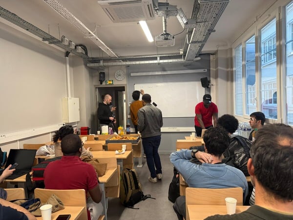 A group of people are gathered in a classroom or workshop setting, with some sitting at desks and others standing near a whiteboard.