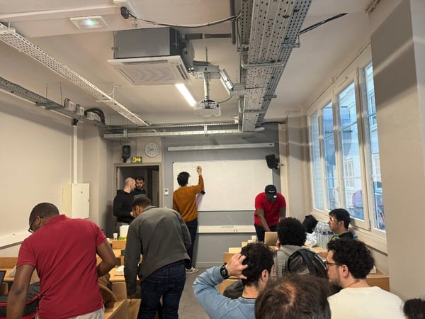 A group of people are gathered in a room with an industrial-style ceiling, with one person writing on a whiteboard while others observe or work at desks.
