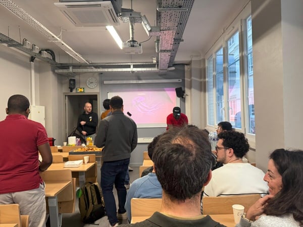 A group of people are gathered in a classroom-style room for a presentation or workshop, with some standing near a table with food and others seated.