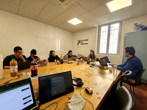 A group of six people are sitting around a large wooden table in an office, working on their laptops with drinks and snacks present.