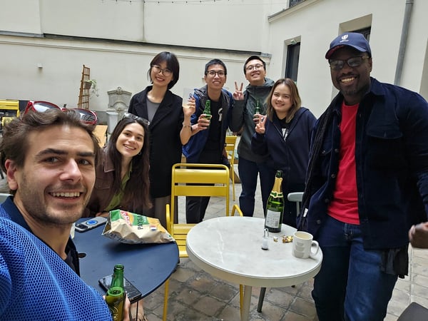 A group of seven diverse young adults are smiling and posing for a photo during a casual outdoor social gathering in a courtyard.