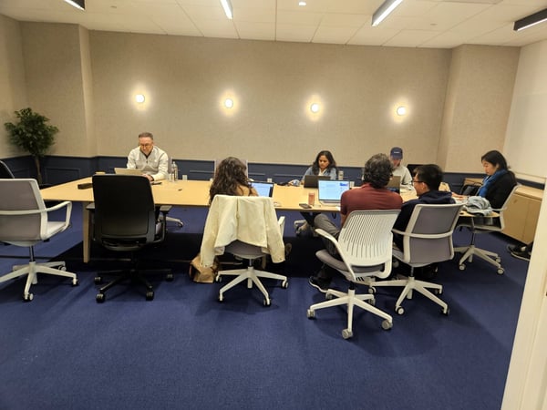 A group of people are seated around a large conference table in a modern office, focused on their laptops.