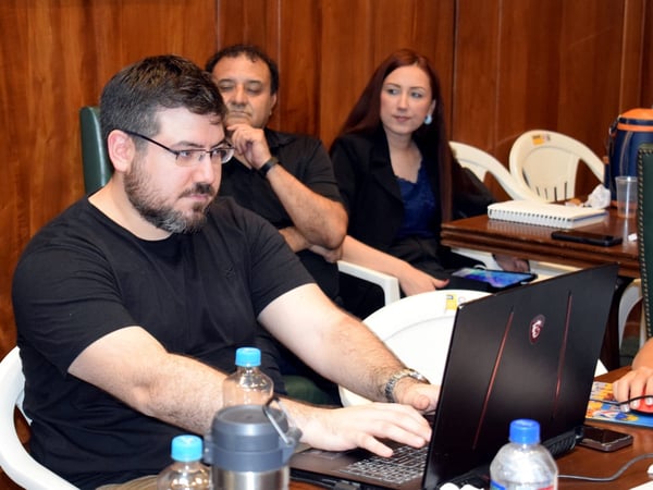 A man is focused on working on a laptop in the foreground, while a man and a woman sit behind him in a room with wooden paneled walls.