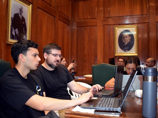 A group of people are focused on their laptops in a room with classic wood-paneled walls and framed portraits.