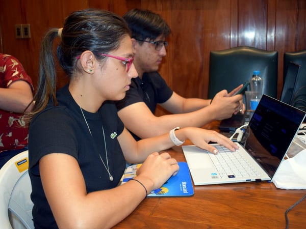 A photograph showing a woman and a man sitting at a wooden table, both focused on their laptops in an indoor room with wood-paneled walls.