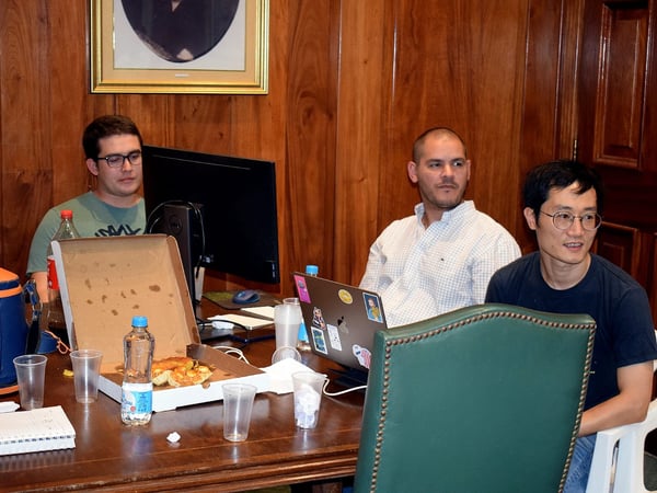 Three men are sitting around a wooden table in a room with wood-paneled walls, appearing to be in the middle of a working session with a pizza box and laptops.