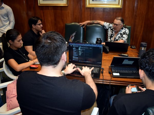 A group of people are gathered around a large wooden table in a wood-paneled room, engaged in a collaborative session with laptops and a smartphone.