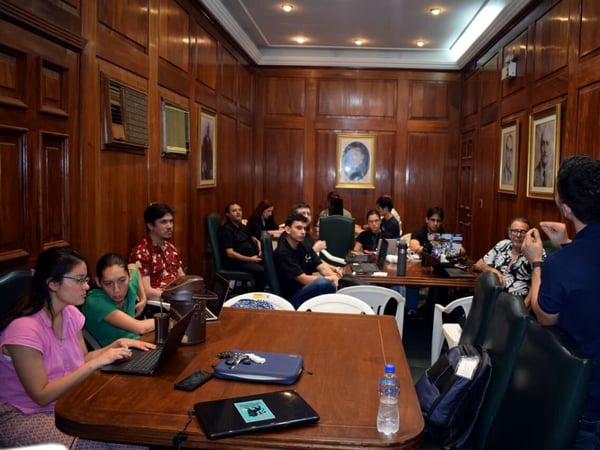A group of people are gathered in a wood-paneled room for a meeting or presentation, with several individuals using laptops while listening to a speaker.