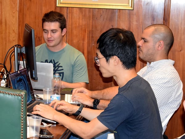 Three men are sitting at a table, focused on their laptops and a computer monitor in a room with wood-paneled walls.