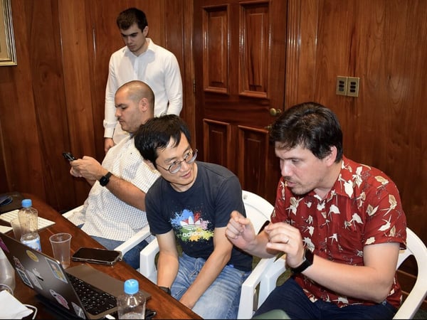 A group of four men are gathered around a table in a wood-paneled room, engaged in a discussion while working on a laptop.