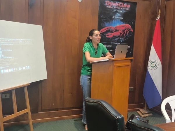 A woman in a green polo shirt stands behind a wooden podium giving a presentation, with a projection screen displaying code to her left and a Paraguayan flag to her right.