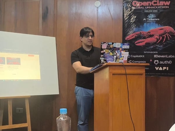 A man stands at a wooden podium giving a presentation, with a projector screen to his left and an event poster on the wall behind him.