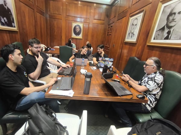 A group of people are gathered around a large wooden table in a wood-paneled room, working on laptops and engaged in discussion.