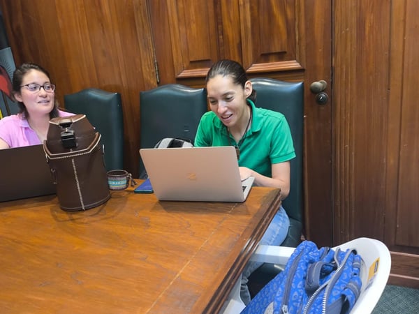 Two women are seated at a large wooden table in a room with wood-paneled walls, both working on laptops.