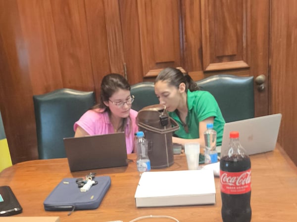 Two women are sitting at a wooden table in a wood-paneled room, focused on a laptop screen together.
