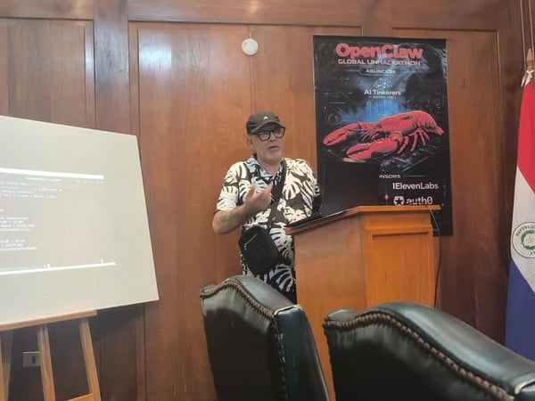 A man in a patterned shirt and cap stands at a wooden lectern giving a presentation. To his left is a screen displaying terminal text, and behind him is a poster for 'OpenClaw Global Unhackathon'.