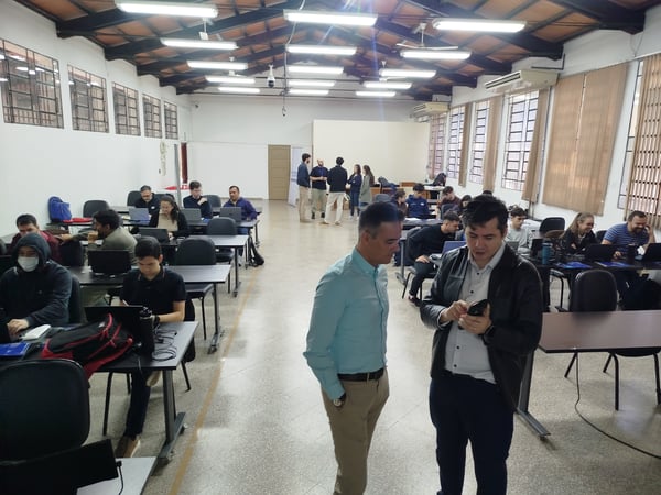 A group of people are gathered in a large, well-lit indoor room with rows of desks, some working on laptops while others stand and talk.