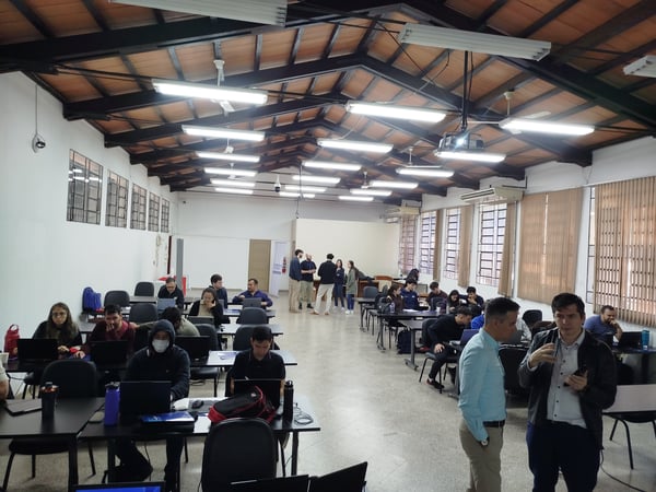A wide-angle photograph of a large indoor workspace or classroom where numerous people are working on laptops at desks, while others stand and converse in the background and foreground.