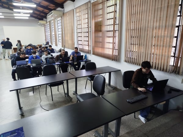 A group of people, mostly young adults, are seated at long tables in a classroom-like setting, working on their laptops during a workshop.