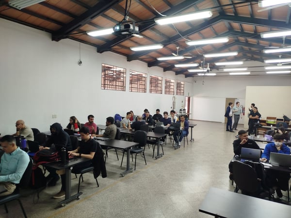 A group of people are working on laptops at long tables in a large, high-ceilinged room with exposed wooden beams and fluorescent lighting.
