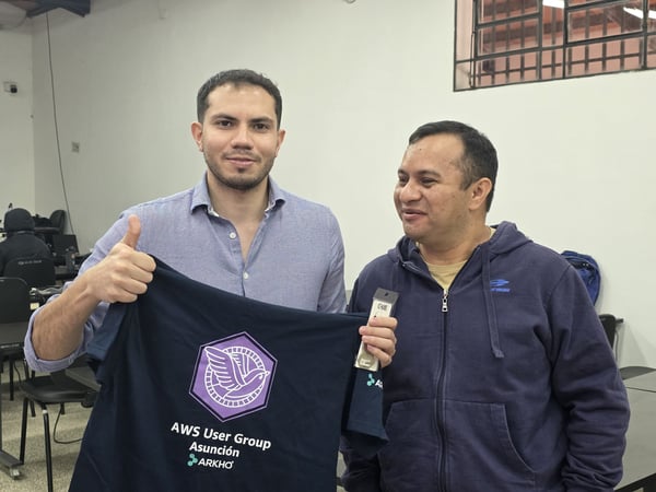 Two men are posing for a photo indoors; one holds up a dark t-shirt with the 'AWS User Group Asunción' logo and gives a thumbs up.
