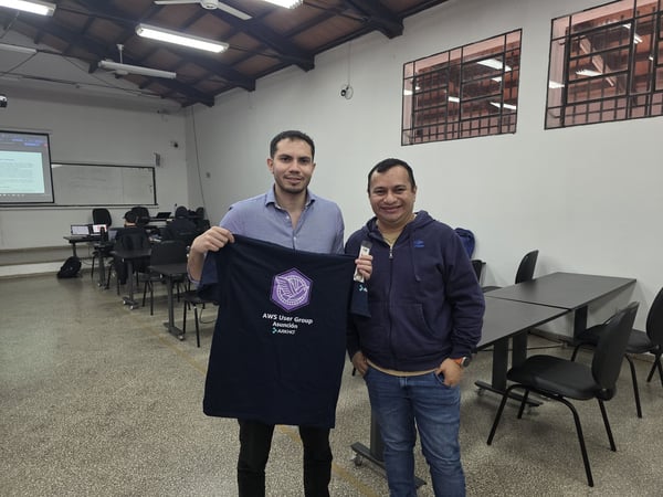 Two men standing in a large indoor room, with one holding up a dark blue t-shirt featuring the AWS User Group Asunción logo.