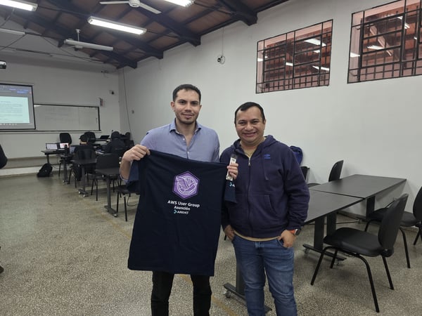 Two men posing in an indoor classroom or office setting, with one holding up a navy blue t-shirt featuring the AWS User Group Asunción logo.
