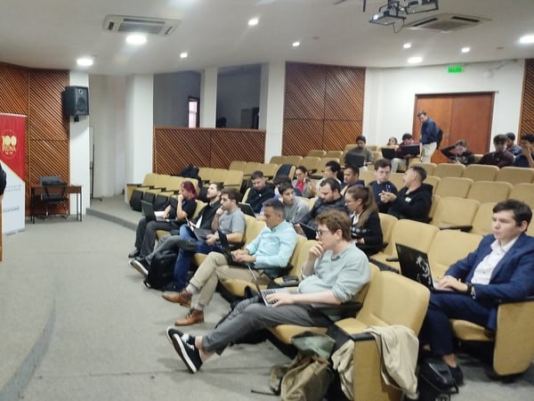 A man stands at the front of an auditorium, giving a presentation to an audience of people seated with their laptops open.