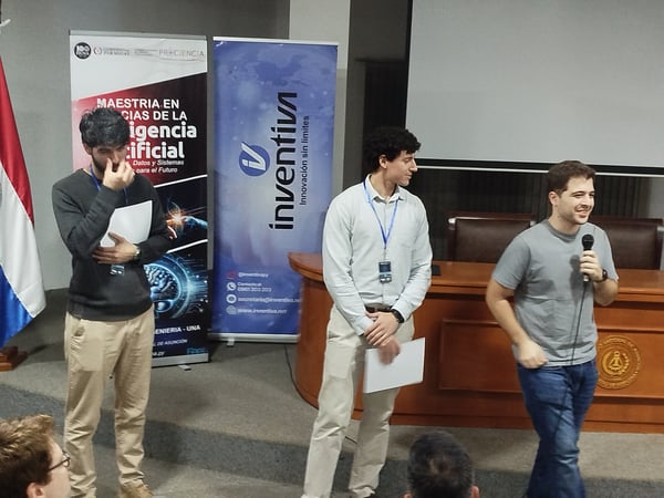 Three men are on a stage during an event, with promotional banners for an Artificial Intelligence master's program and a company named Inventiva in the background.