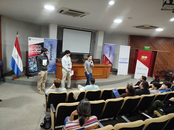 Three men are giving a presentation on a stage in an auditorium with an audience seated in the foreground and several promotional banners in the background.